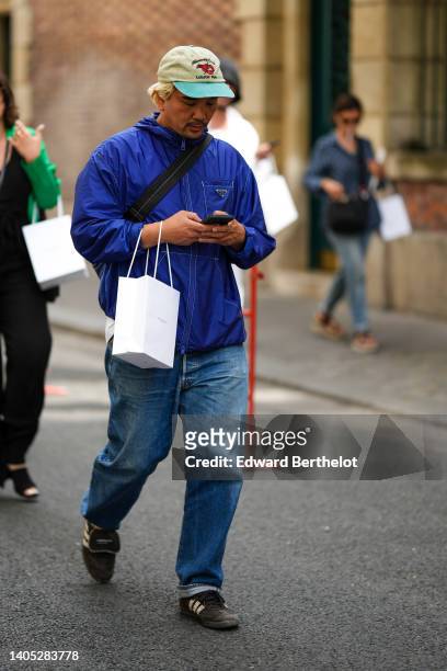 Guest wears a beige and blue with embroidered red pattern cap, a royal blue nylon jacket from Prada, blue faded denim large pants, black suede with...