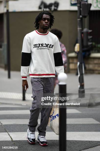 Guest wears white with black and red print pattern sweater, gray cargo pants, white leather with black varnished sneakers from Jordan, outside the...