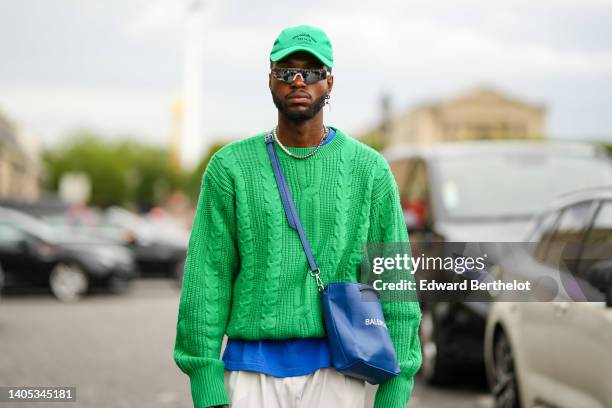 Guest wears a green with black embroidered inscription pattern cap, black futurist sunglasses, silver earrings, a white and silver pearls necklace, a...