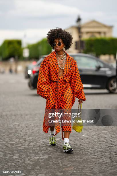 Sarah Monteil wears black sunglasses, oversized silver earrings, silver large chain necklaces, a navy blue with orange print pattern oversized silk...