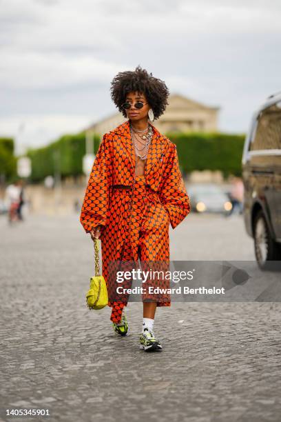 Sarah Monteil wears black sunglasses, oversized silver earrings, silver large chain necklaces, a navy blue with orange print pattern oversized silk...