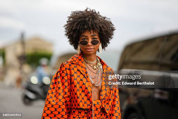 Sarah Monteil wears black sunglasses, oversized silver earrings, silver large chain necklaces, a navy blue with orange print pattern oversized silk...