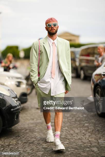 Karb wears blue sunglasses, a white shirt, a pale pink tie, a pale green shoulder pads oversized blazer jacket, matching pale green suit shorts, a...