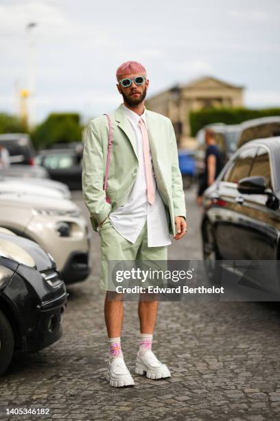 Karb wears blue sunglasses, a white shirt, a pale pink tie, a pale green shoulder pads oversized blazer jacket, matching pale green suit shorts, a...