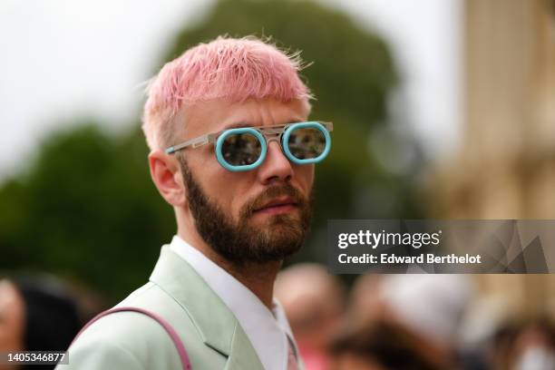 Karb wears blue sunglasses, a white shirt, a pale pink tie, a pale green shoulder pads oversized blazer jacket, a pink shiny leather shoulder bag,...