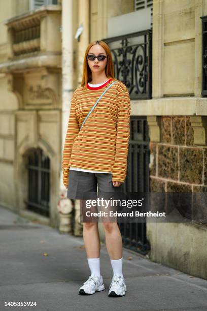 Model wears black sunglasses, a gold chain necklace, a white t-shirt, a red / orange / yellow striped print pattern long sleeves sweater, a pale blue...