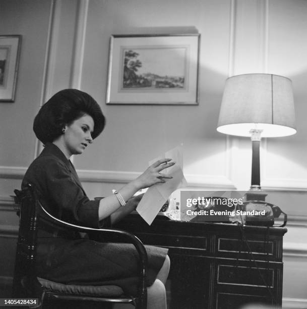 British socialite Lady Elizabeth Shakerley , sitting at a desk on which are two telephones as she reads a document with a cigarette held between her...
