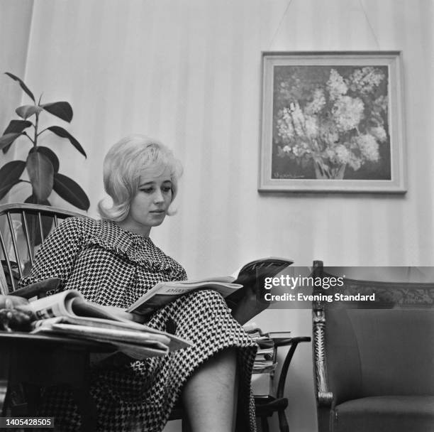 British socialite and actress Elizabeth Rees-Williams wearing a dogtooth check outfit as she sits in a chair reading a magazine, a framed image of...