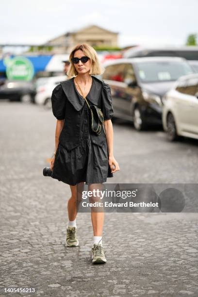 Guest wears black sunglasses, a gold chain pendant cross necklace, a black puffy short sleeves / ruffled buttoned shirt dress, a black with...