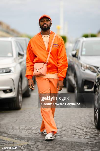 Guest wears an orange cap from Converse, black glasses, a white and silver pearls necklace, a pale orange embroidered sweater from Thom Browne, a...