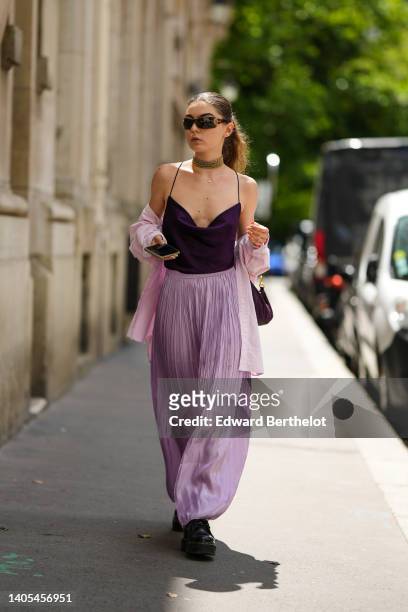 Guest wears futurist sunglasses, gold earrings, a large gold necklace, a gold chain pendant necklace, a dark purple satin V-neck tank-top, a pale...