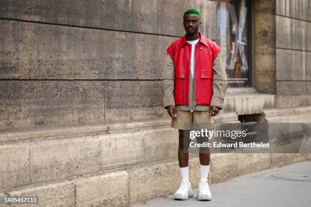 Guest wears a white t-shirt, a silver chain necklace, a red shiny leather sleeveless jacket, a beige cargo jacket, beige suit pants, white socks,...