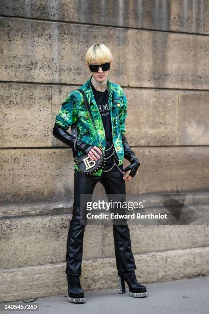 Guest wears black futurist sunglasses from Versace, a rhinestones large necklace, a black with silver inscription print pattern t-shirt from Balmain,...
