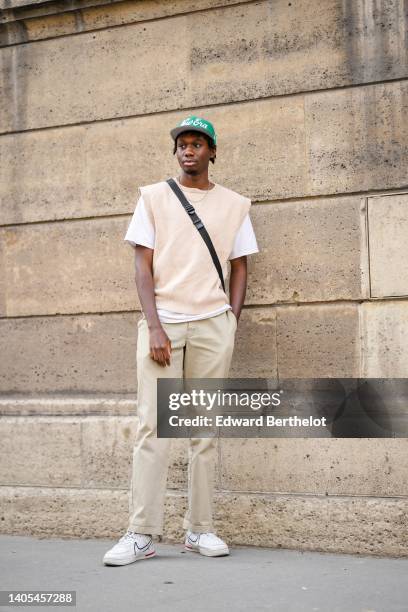 Guest wears a green with white inscriptions pattern cap, a silver chain necklace, a black crossbody bag, a white t-shirt, a beige sleeveless wool...