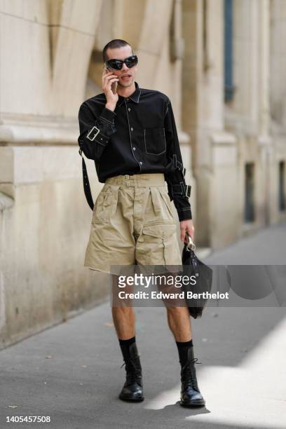 Guest wears sunglasses, a black denim shirt, beige cargo shorts, a black shiny leather handbag, black socks, black shiny leather laces ankle boots,...