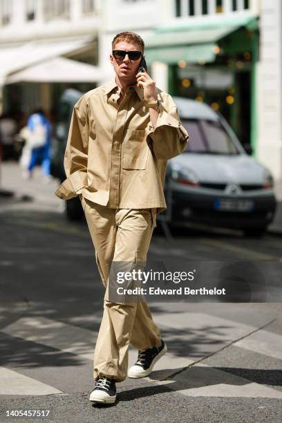 Guest wears black sunglasses, a beige oversized shirt, matching beige cargo pants, black laces ankle sneakers from Converse All Star, a gold watch ,...