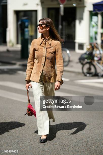 Guest wears black sunglasses, a brown suede shirt, white striped print pattern wide legs pants, black shiny varnished leather ballerinas, a camel...