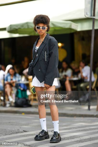 Guest wears white sunglasses, gold pendant and rhinestones earrings, a black asymmetric blazer jacket, a white square-neck / pleated short dress,...