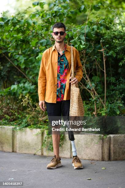 Guest wears sunglasses, a gold chain necklace, an orange shirt, a black with red / green flower print pattern shirt, a beige oversized wicker...