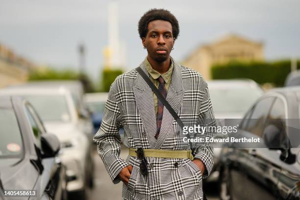 Guest wears silver earrings, a khaki shirt, a gold chain necklace, a brown and black checkered print pattern silk tie, a black and white houndstooth...