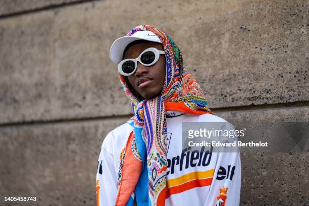 Guest wears a white cap, white sunglasses, a multicolored print pattern scarf, a white with red and orange print pattern sweater, outside the Wooyoun...