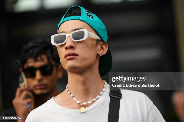 Model wears a neon blue with black embroidered pattern cap, white sunglasses, a white pearls and gold necklace, a white t-shirt, a black crossbody...