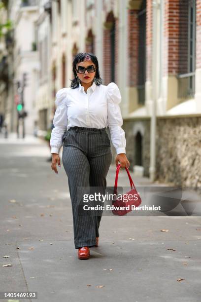 Guest wears black large sunglasses, silver pendant earrings, a white puffy sleeves / ruffled shirt, black striped print pattern large pants, a red...