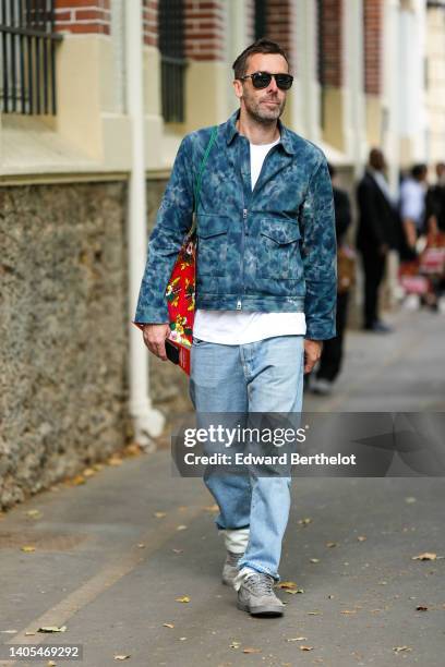 Guest wears black sunglasses, a white t-shirt, a blue tie and dye print pattern zipper jacket, a red with multicolored flower print patter shoulder...