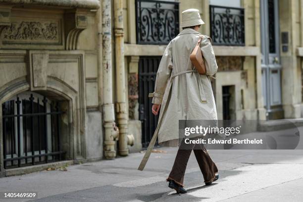 Guest wears a beige bob hat, a white latte ribbed zipper high neck cropped polo shirt, a beige long trench coat, a brown shiny leather shoulder bag,...