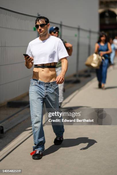 Guest wears black sunglasses, a white cropped t-shirt, a black corset top, blue denim large pants, black shiny suede sneakers from Off-White, outside...