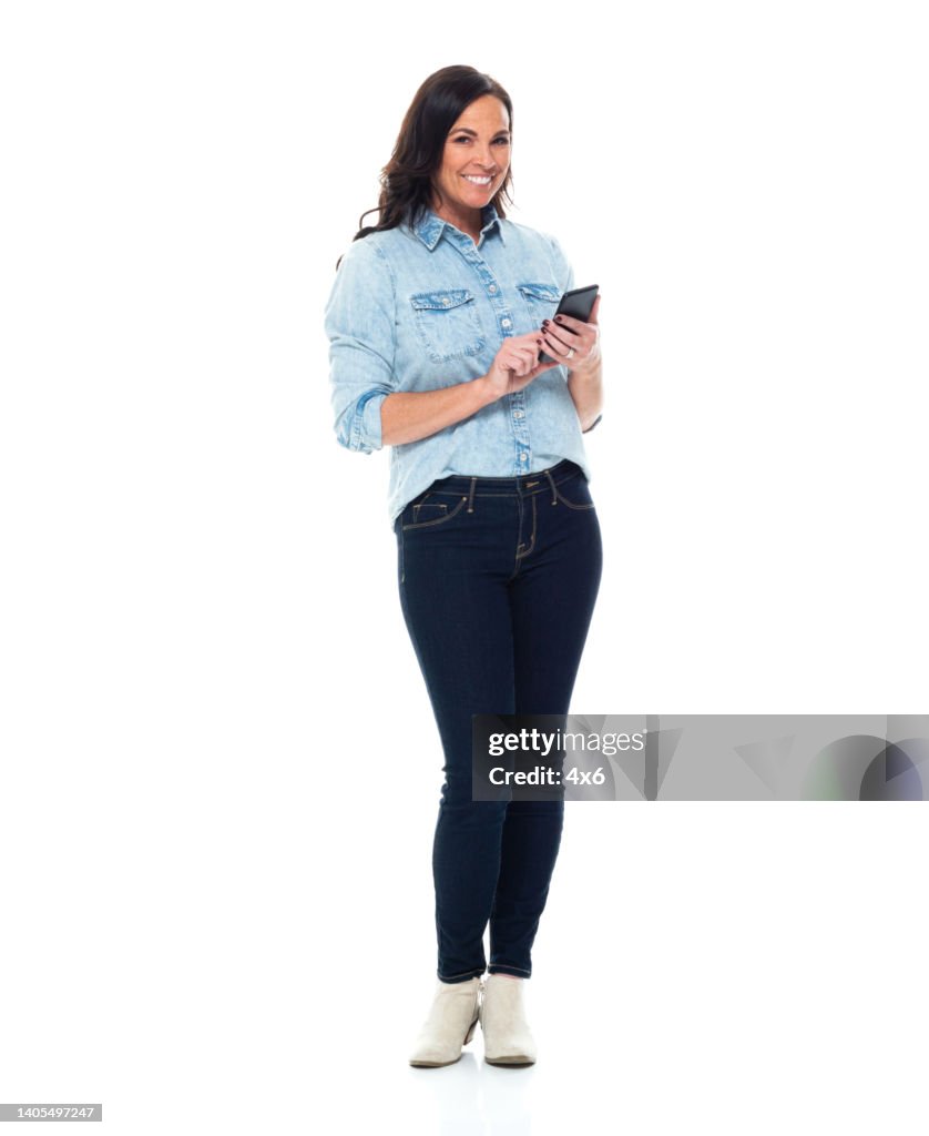 Caucasian female standing in front of white background wearing jeans and using smart phone