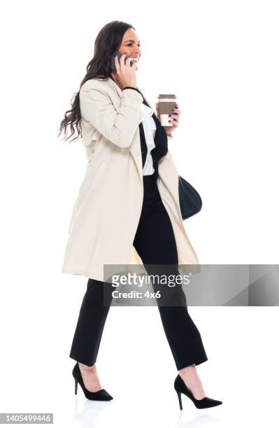 caucasian young women manager walking in front of white background wearing businesswear and holding purse and using mobile phone - business woman side stockfoto's en -beelden