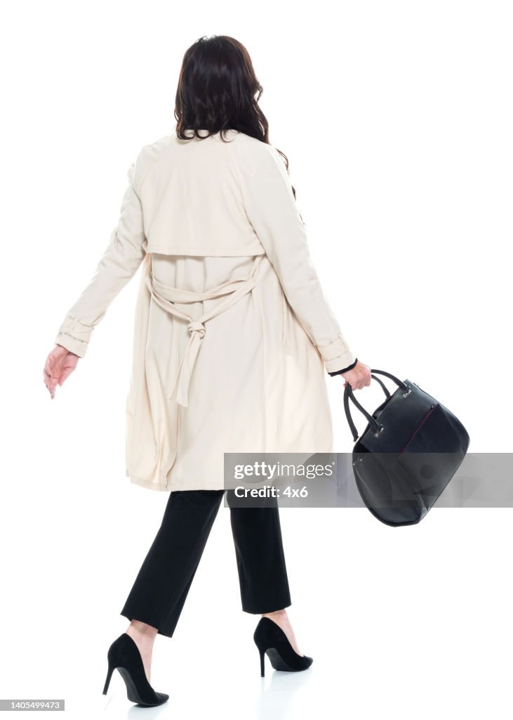 Caucasian young women businesswoman walking in front of white background wearing trench coat and holding purse