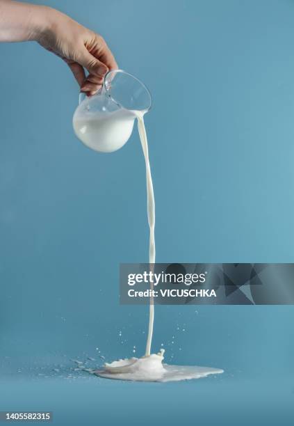 women hand pouring milk from transparent glass jug with splashing at blue background. liquid in motion. - goot stockfoto's en -beelden