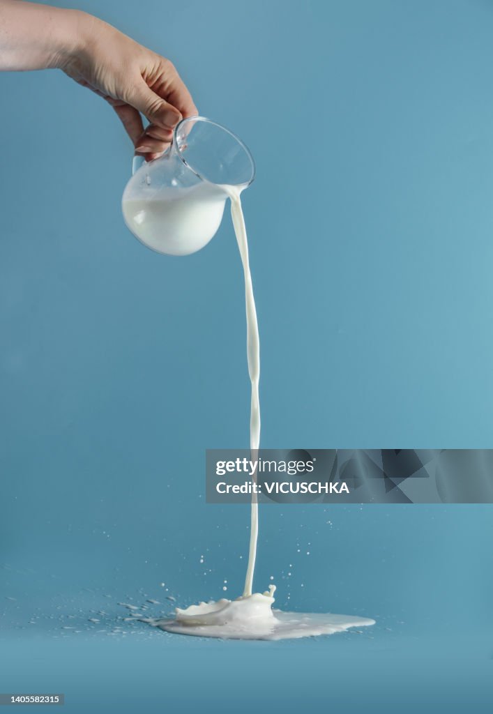 Women hand pouring milk from transparent glass jug with splashing at blue background. Liquid in motion.