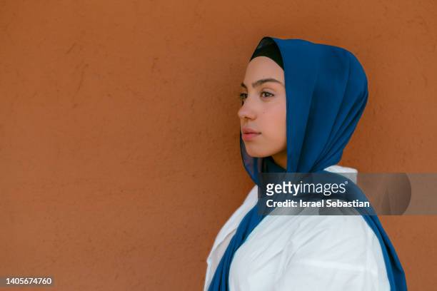 portrait of a young arab woman wearing a blue hijab looking to the side on a brown background. - teal portrait stockfoto's en -beelden