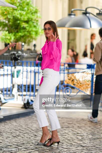 Nieves Alvarez wears brown sunglasses, a neon pink knot necklace silk shirt, white buttoned high waist denim cigarette jeans pants, a neon pink shiny...