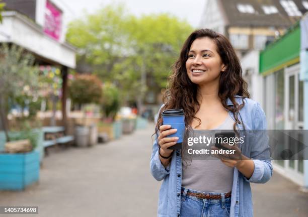 happy woman drinking a cup of coffee on the move while using her phone - take away coffee cup stock pictures, royalty-free photos & images