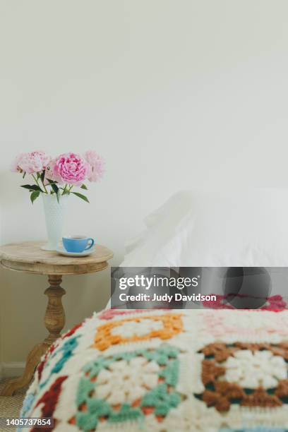 bed with granny square colorful afghan and side table with vase of pink peonies and blue cup of tea - arte de la costura fotografías e imágenes de stock