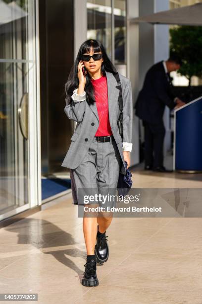 Guest wears black sunglasses, a gold ring, a red cut-out striped pattern pullover, a gray checkered print pattern long blazer jacket, matching gray...