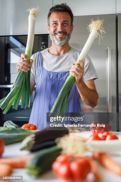 smiling mature man with beard in front of a table full of fresh vegetables holding two leeks. healthy food concept. - ingredientes cocina stock-fotos und bilder