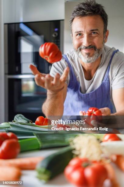 smiling 50 year old man with a beard in front of a table full of fresh vegetables tossing a tomato. healthy food concept. - ingredientes cocina stock-fotos und bilder