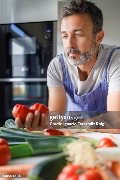 bearded mature man in front of a table full of fresh vegetables looking at some tomatoes.healthy food concept. - ingredientes cocina stock-fotos und bilder