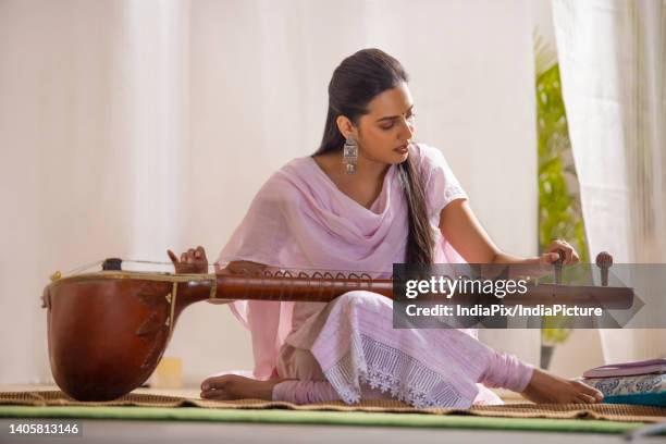 young female musician tuning her sitar while sitting in living room - sitar stock stock pictures, royalty-free photos & images