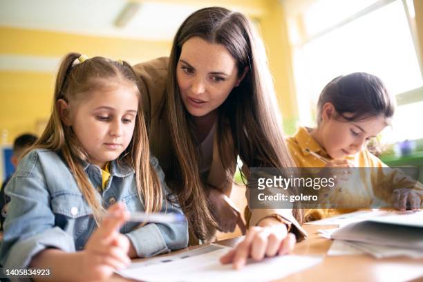 junge lehrerin erklärt schulmädchen am schreibtisch im unterricht - elementary school building stock-fotos und bilder