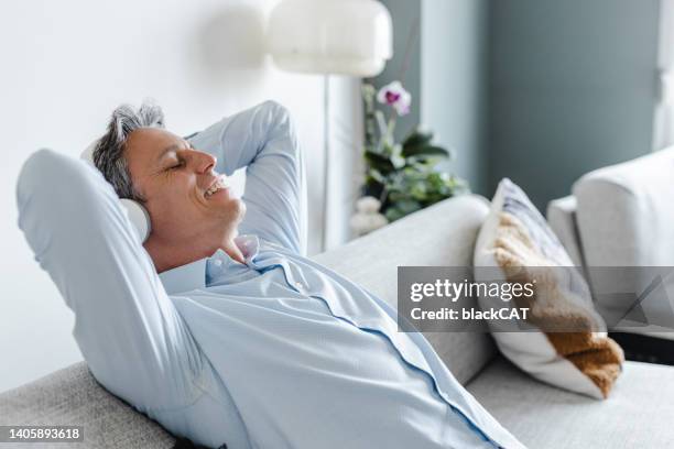 mature man with hands behind head, relaxing on sofa and listening to music with headphones in the living room - white sofa stock pictures, royalty-free photos & images