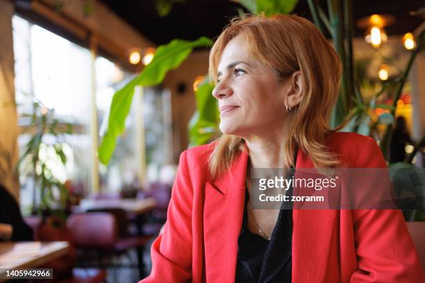 elegante frau in roter jacke, die in der stadtbar sitzt und mit einem lächeln aus dem fenster schaut - albanian woman stock-fotos und bilder