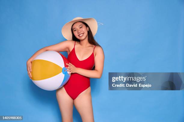 smiling asian woman wearing a swimwear and a hat while holding a beach ball over an isolated background. - asian woman swimsuit stock-fotos und bilder