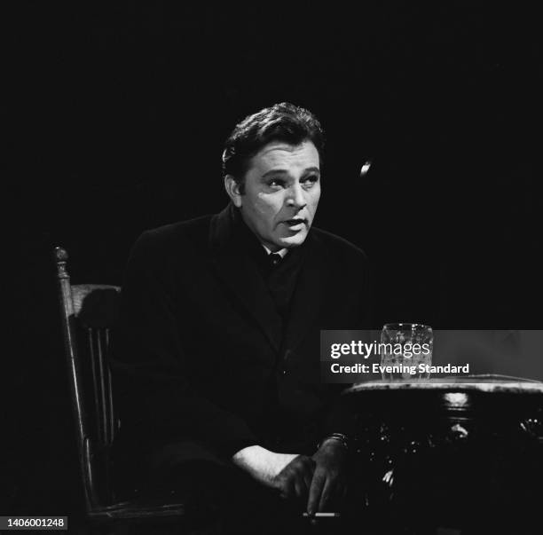 British actor Richard Burton , wearing a black coat and sitting at a table on which is a pint of beer, during an event, United Kingdom, 8th April...