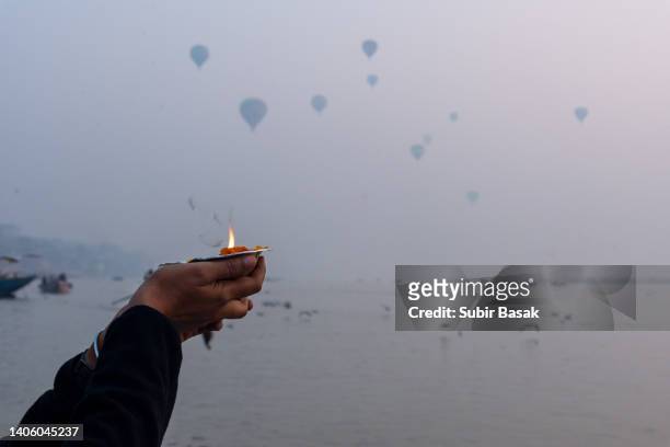 woman praying with oil lamp on the bank of river ganges in the morning with hot air balloons in background ,varanasi,uttar pradesh,india. - diya oil lamp stockfoto's en -beelden